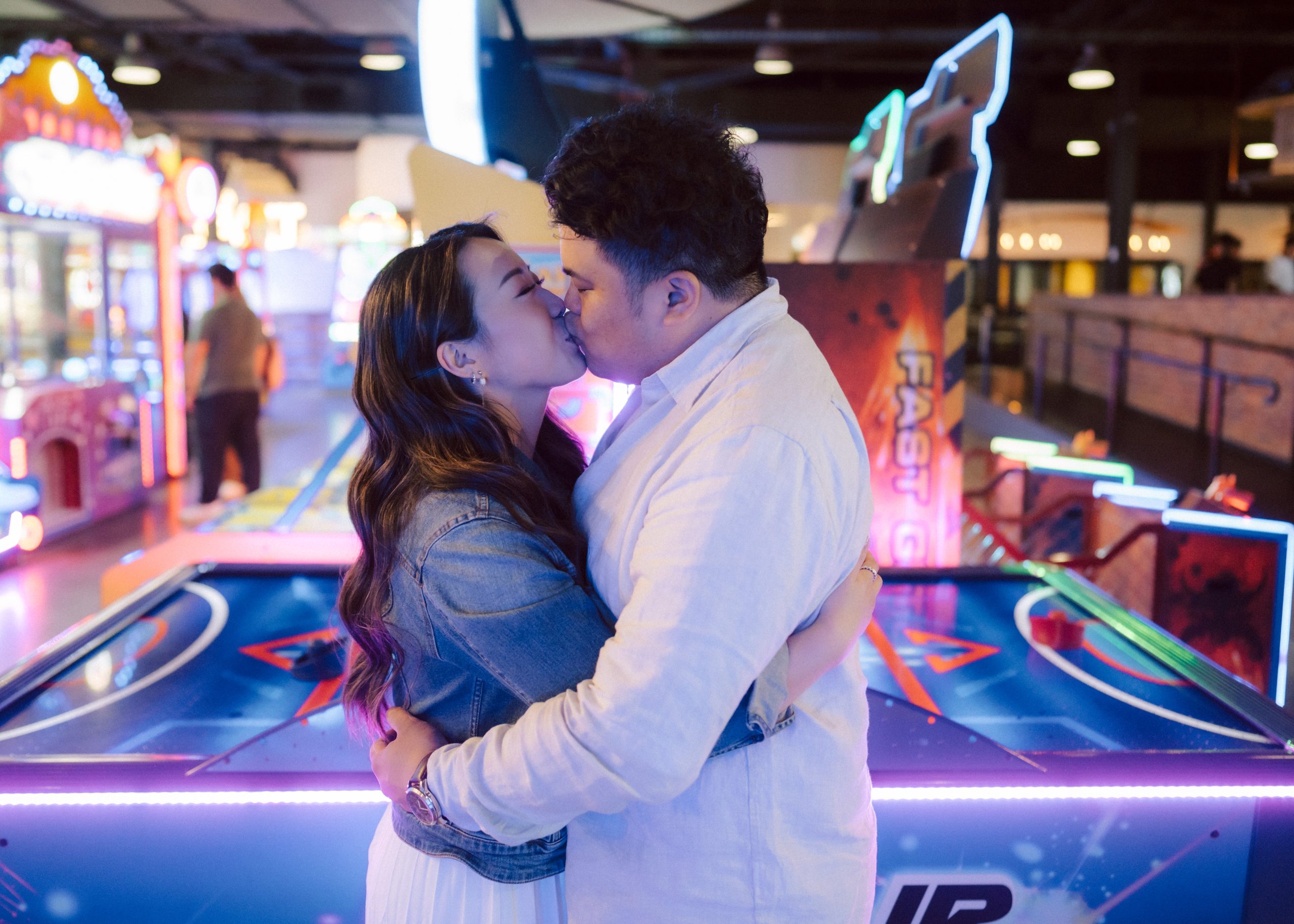 Couple in an embrace at an arcade. Arcade has neon lights in blues and pinks for a pre wedding photoshoot.